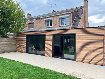 Photographie d'une extension de maison avec bardage bois clair, terrasse carrelée devançant une pelouse